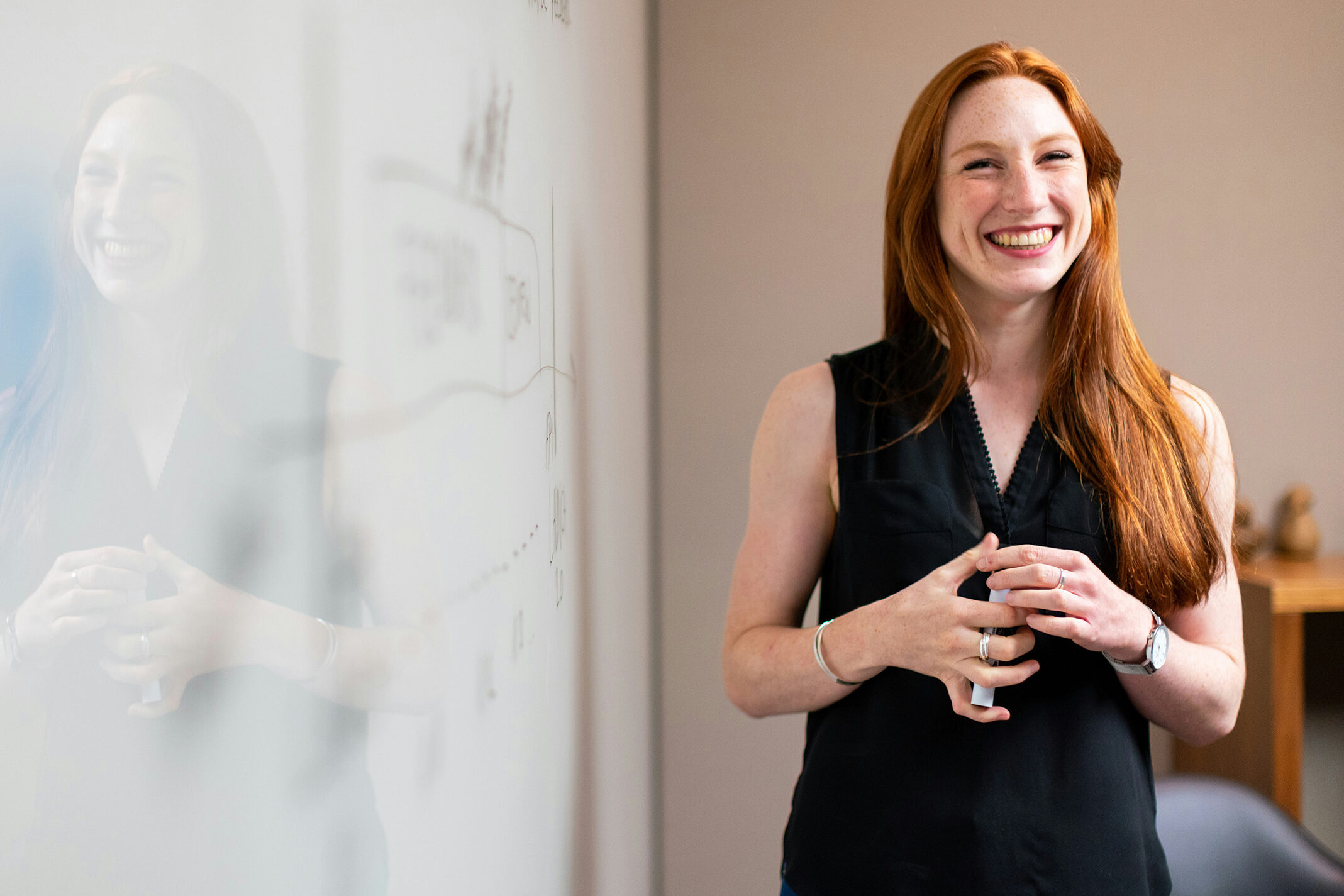 Eine rothaarige Frau lächelt und steht neben einer Whiteboard-Tafel mit einer Zeichnung darauf.
